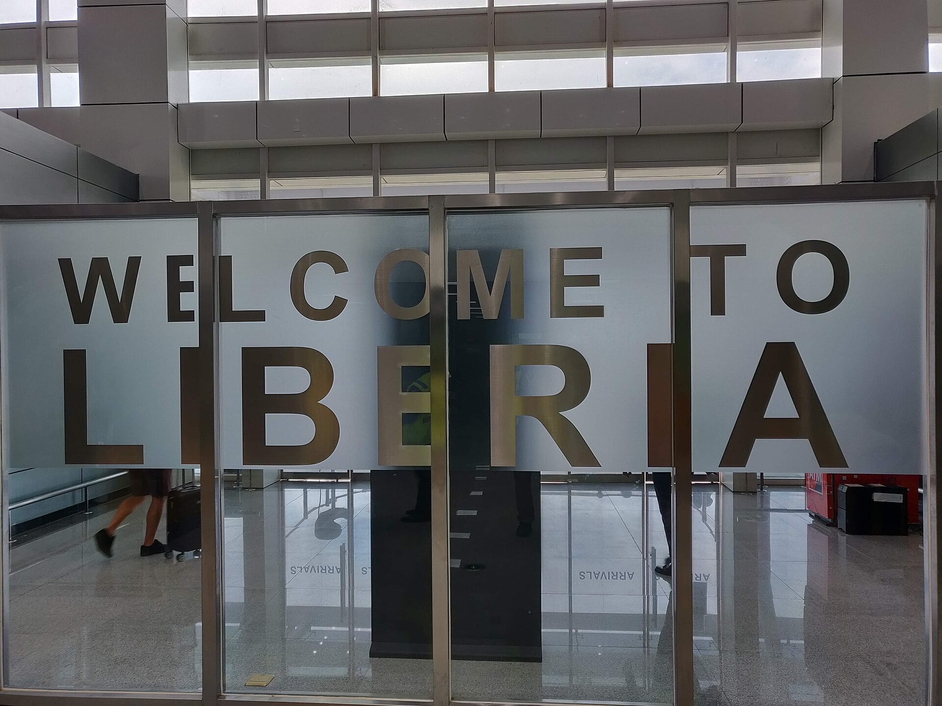 Frosted glass doors with the words Welcome to Liberia in large letters in the arrivals area of Roberts International Airport, with travellers' legs and a suitcase visible behind