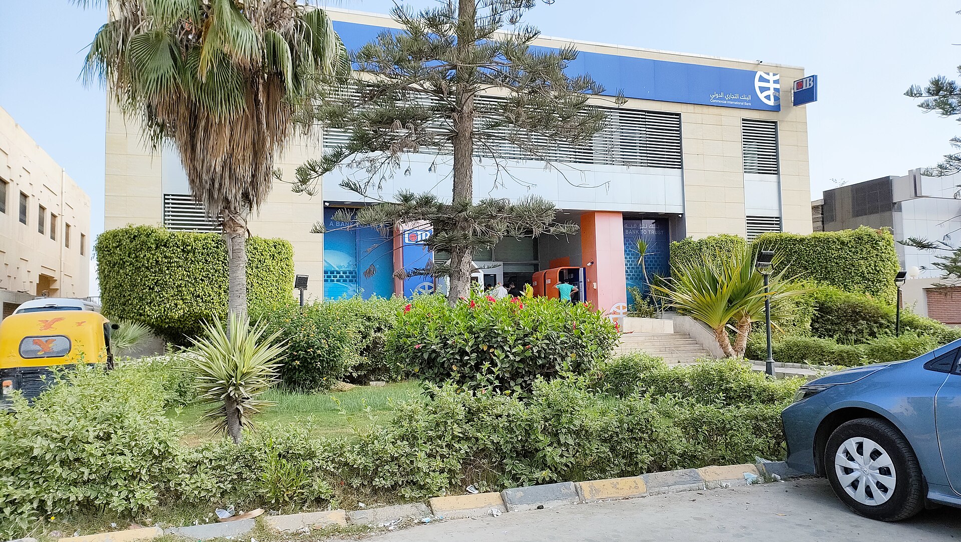 A Commercial International Bank branch with a blue sign behind palms and hedges, a yellow tuk-tuk and a parked car in front
