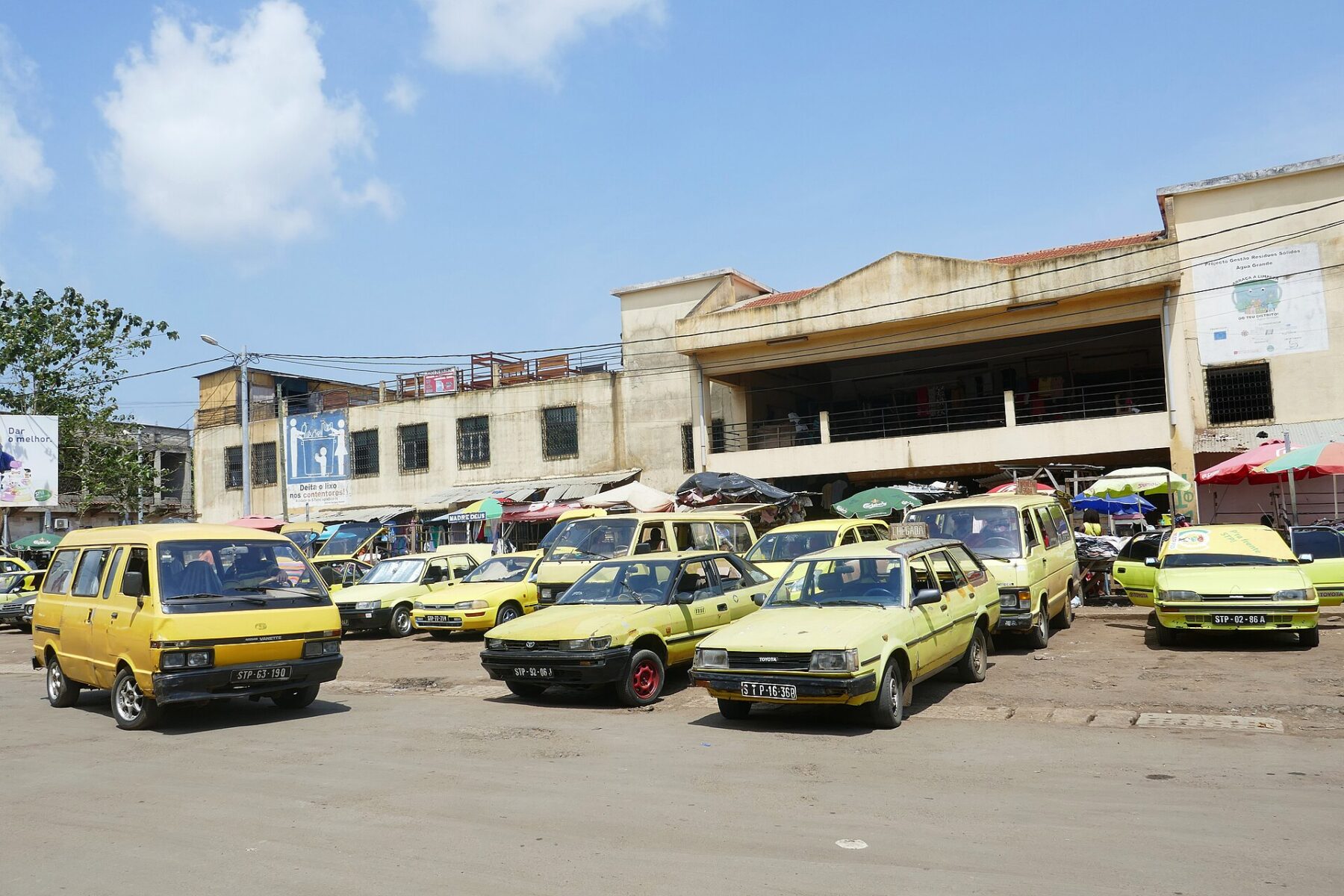 Yellow taxis and minibuses parked in front of a two-storey market building in São Tomé under a blue sky