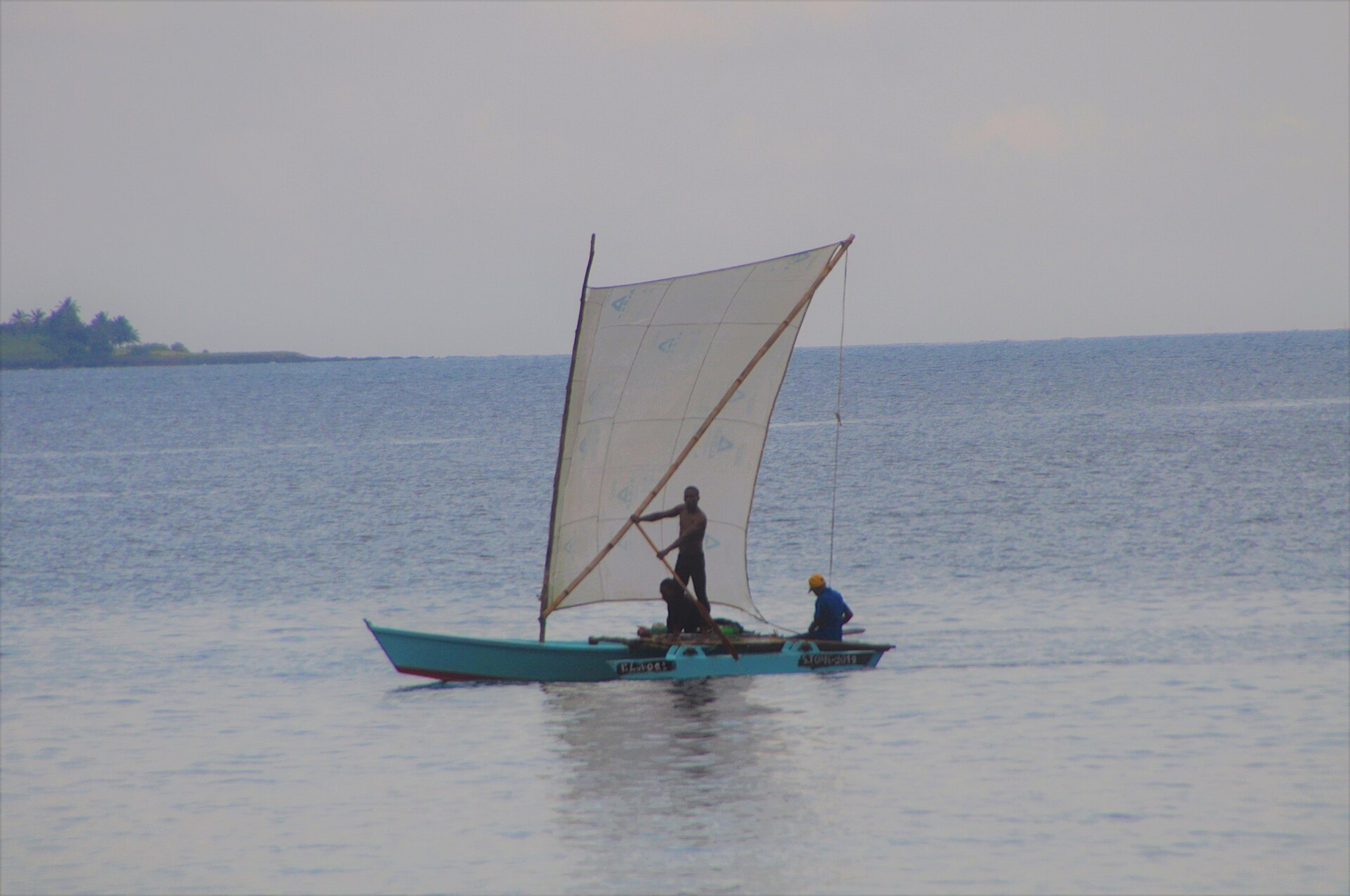 Three fishermen on a turquoise outrigger canoe with a homemade white sail on calm sea off São Tomé