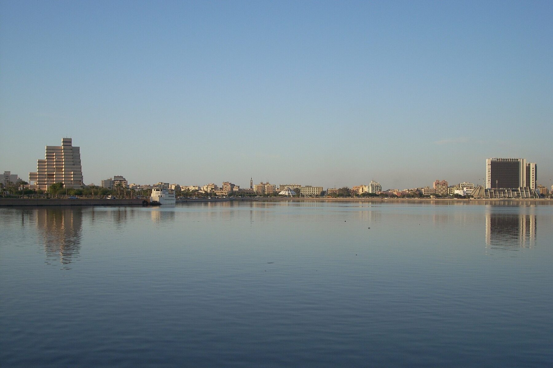 The Benghazi skyline seen across a calm lake under a clear blue sky, with tall buildings at left and right