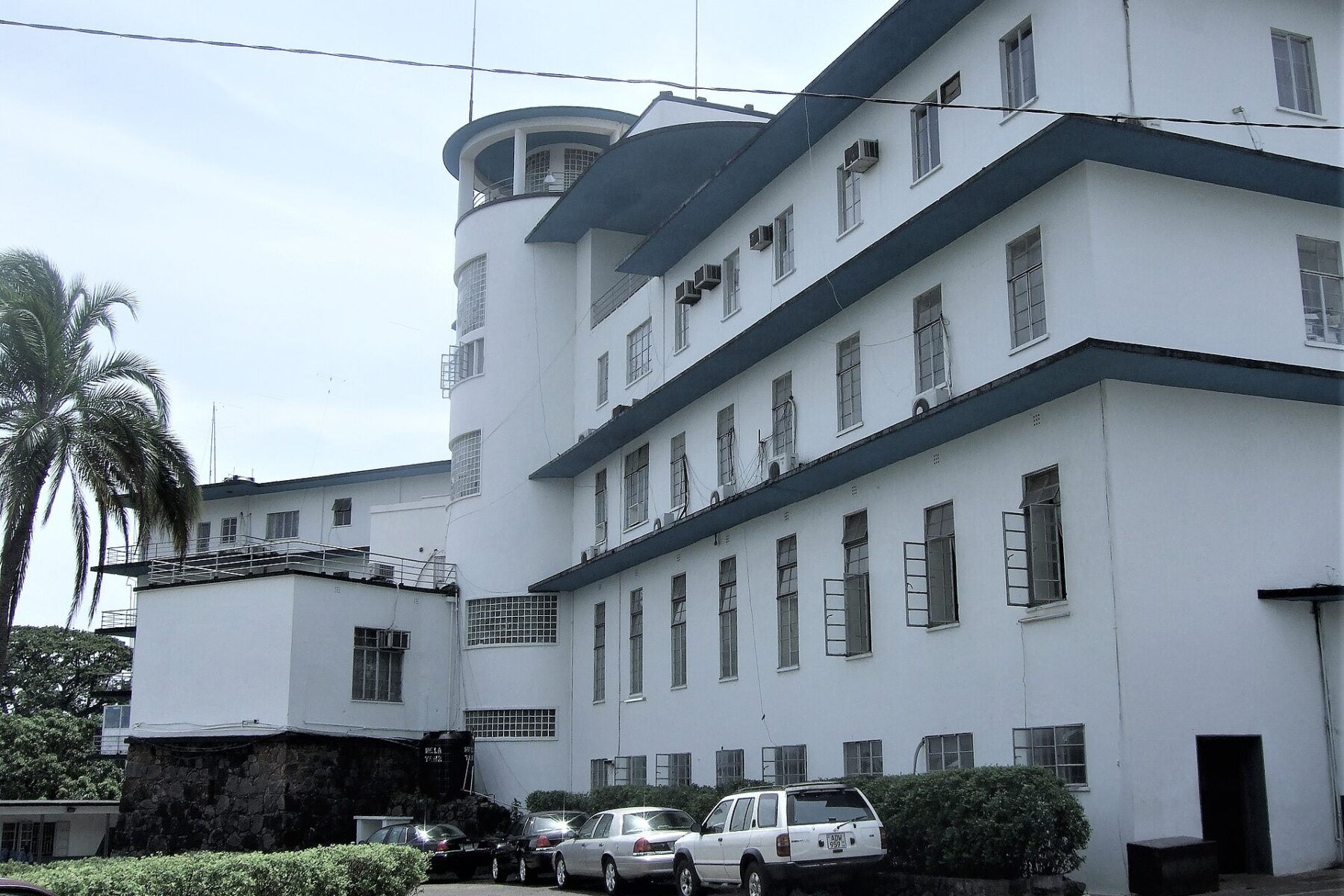 White multi-storey State House building with a round tower, a palm tree and parked cars in front