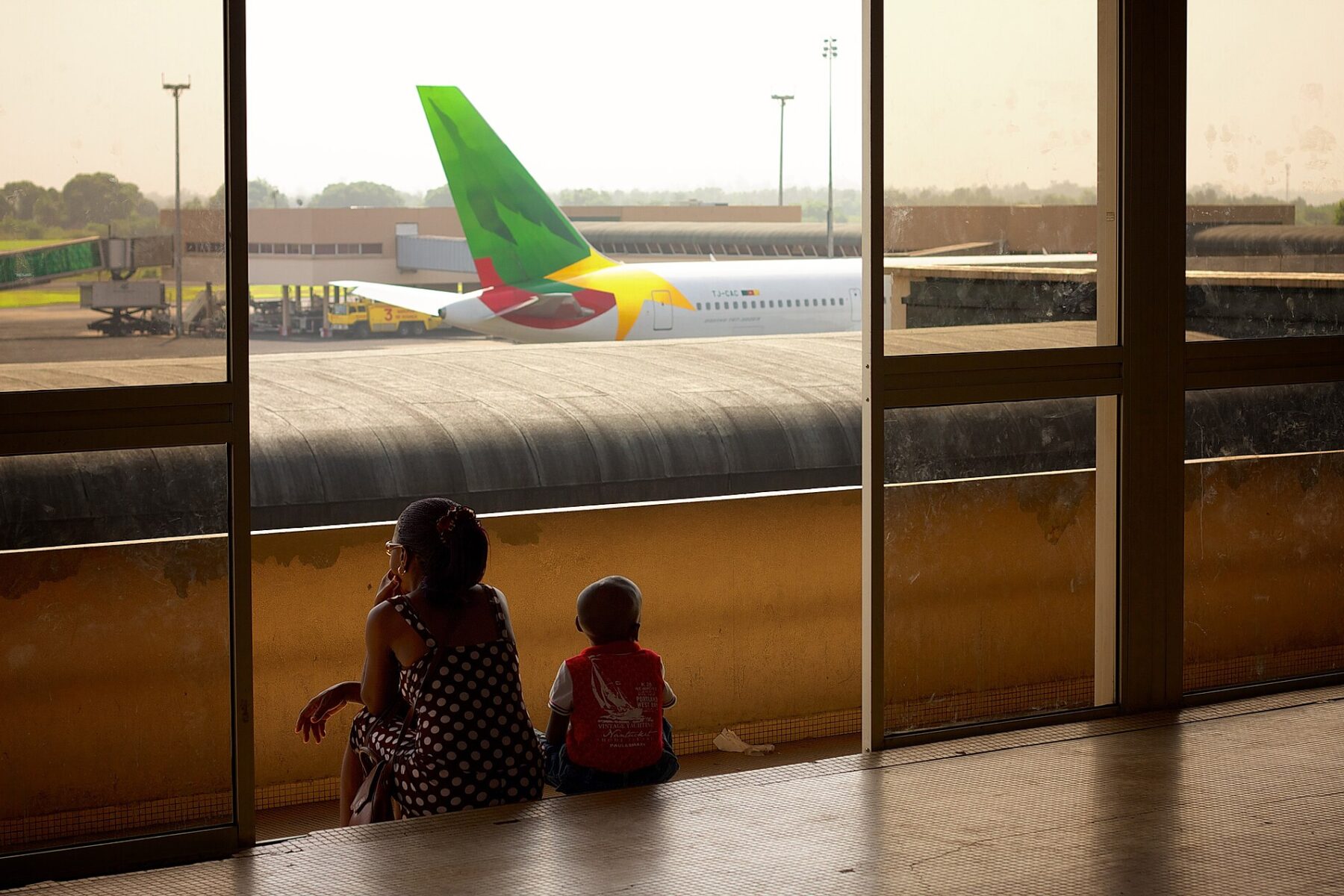 A woman and a small child sit by a terminal window watching a Cameroon Airlines jet parked on the apron at Douala airport