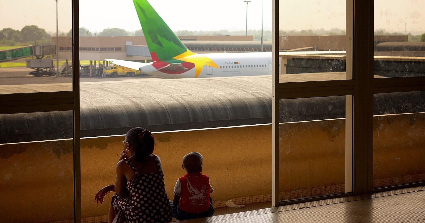 A woman and a small child sit by a terminal window watching a Cameroon Airlines jet parked on the apron at Douala airport