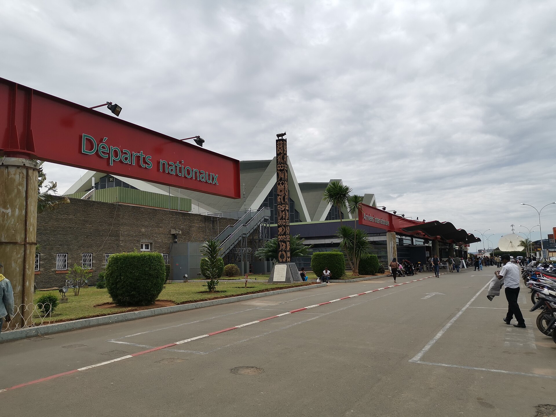 Red departures sign and terminal building of Ivato International Airport in Antananarivo with a road, palm trees and parked motorbikes under a cloudy sky