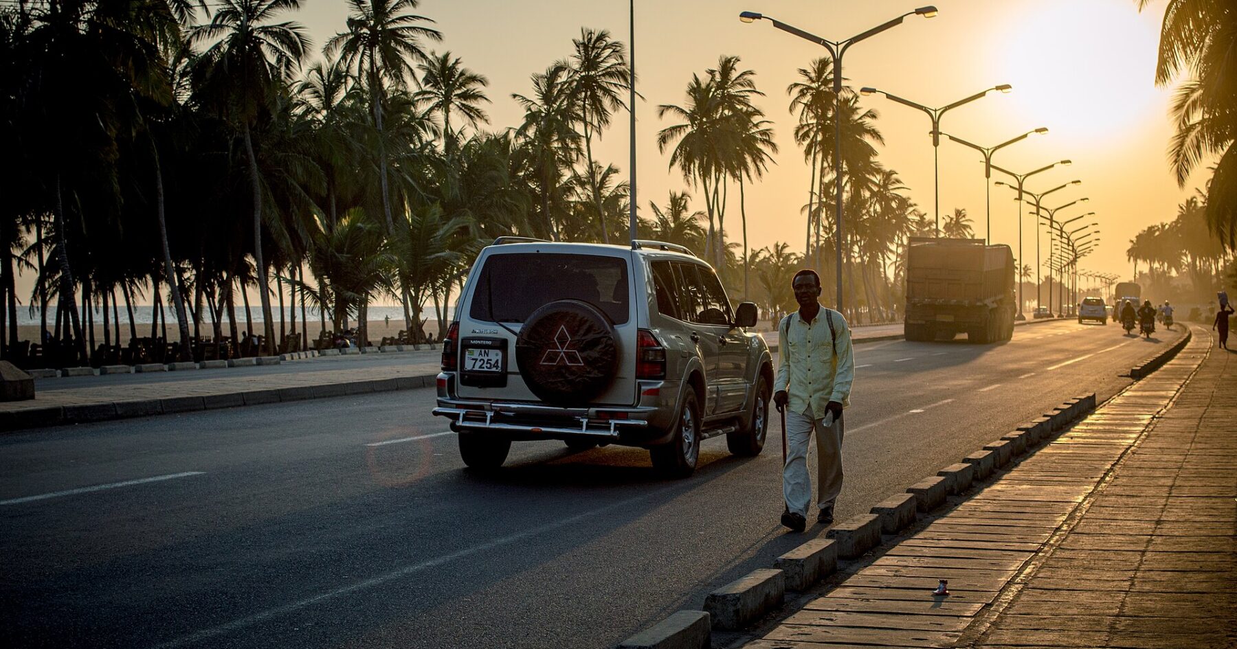 A man walks along the palm-lined coastal road in Lomé at sunset as an SUV and a truck pass