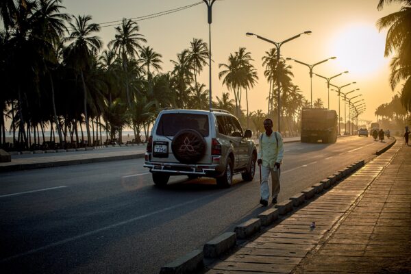 A man walks along the palm-lined coastal road in Lomé at sunset as an SUV and a truck pass