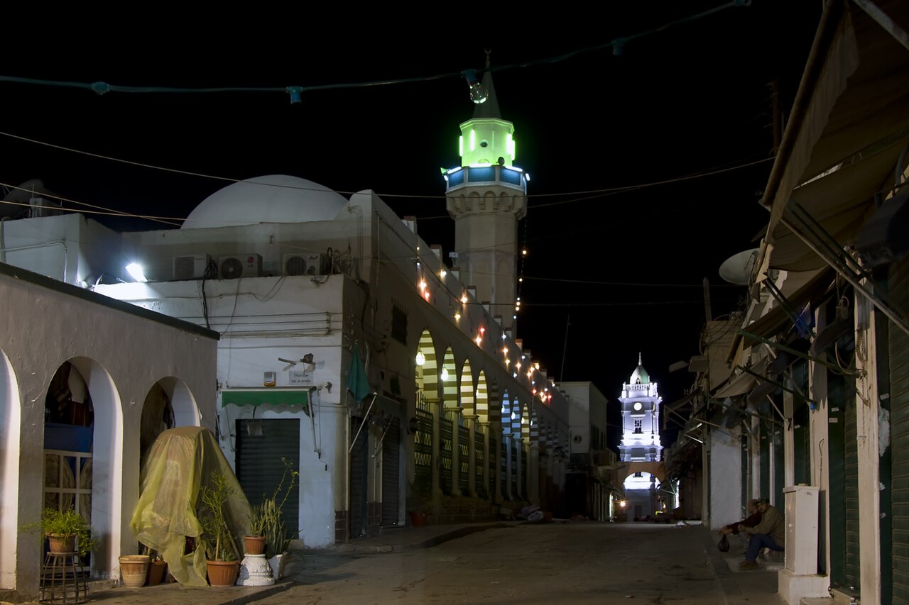 A narrow street in Tripoli's old city at night, with a lit minaret and a clock tower at the end and shuttered shops on the right