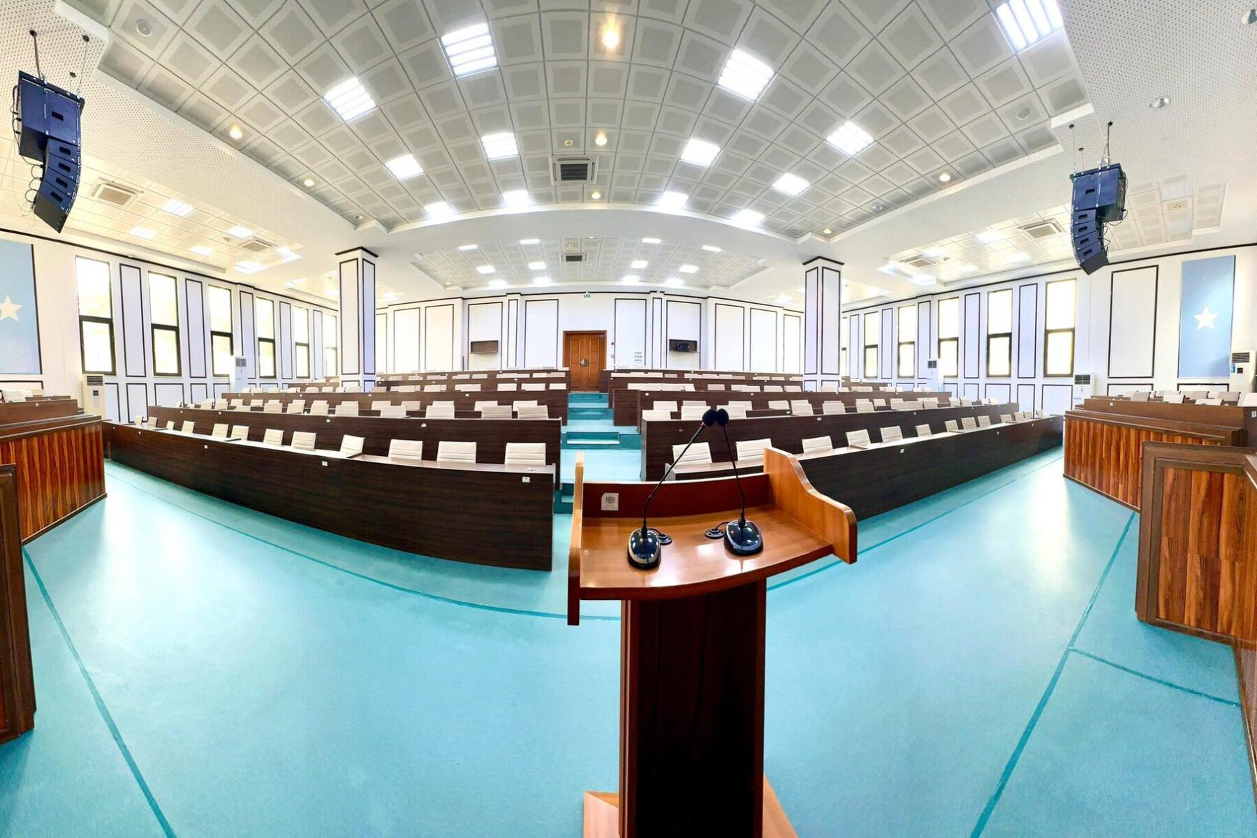 Empty parliamentary chamber with curved wooden benches, a lectern with microphones and blue banners with a white star