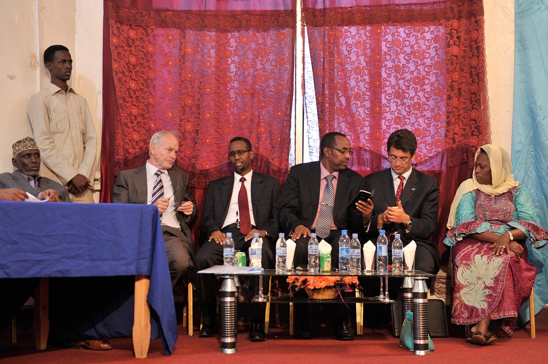 Six people in suits and traditional dress seated behind water bottles in front of red curtains at a signing ceremony in Mogadishu