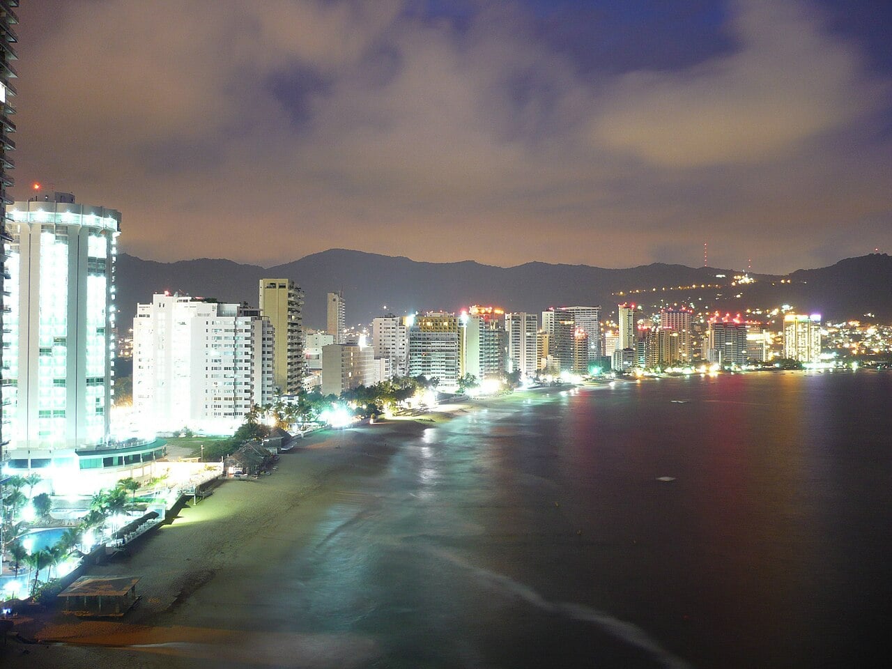 Hotel towers along the beach of Acapulco Bay at night, with mountains behind.
