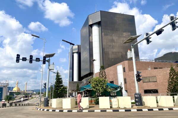 Dark glass office tower with white columns beside a road junction in Abuja, Nigeria, under a blue sky with clouds