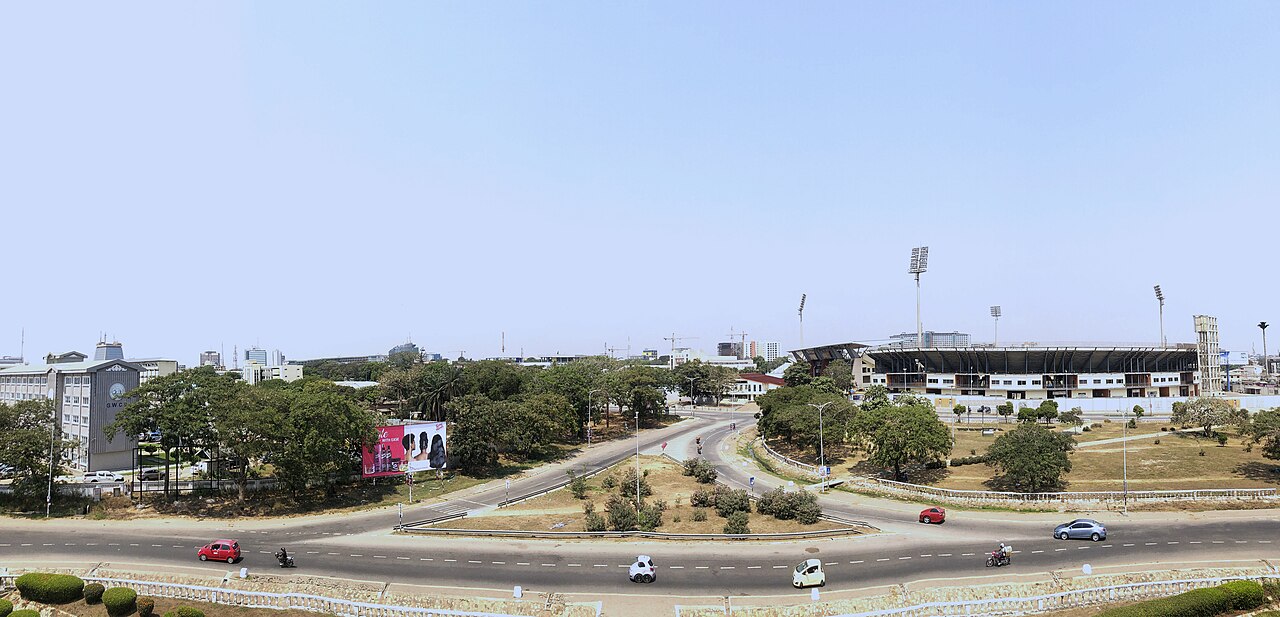 Panoramic view over a road roundabout in Accra, Ghana, with Black Star Square and the Ohene Djan Sports Stadium floodlights on the right under a clear sky.