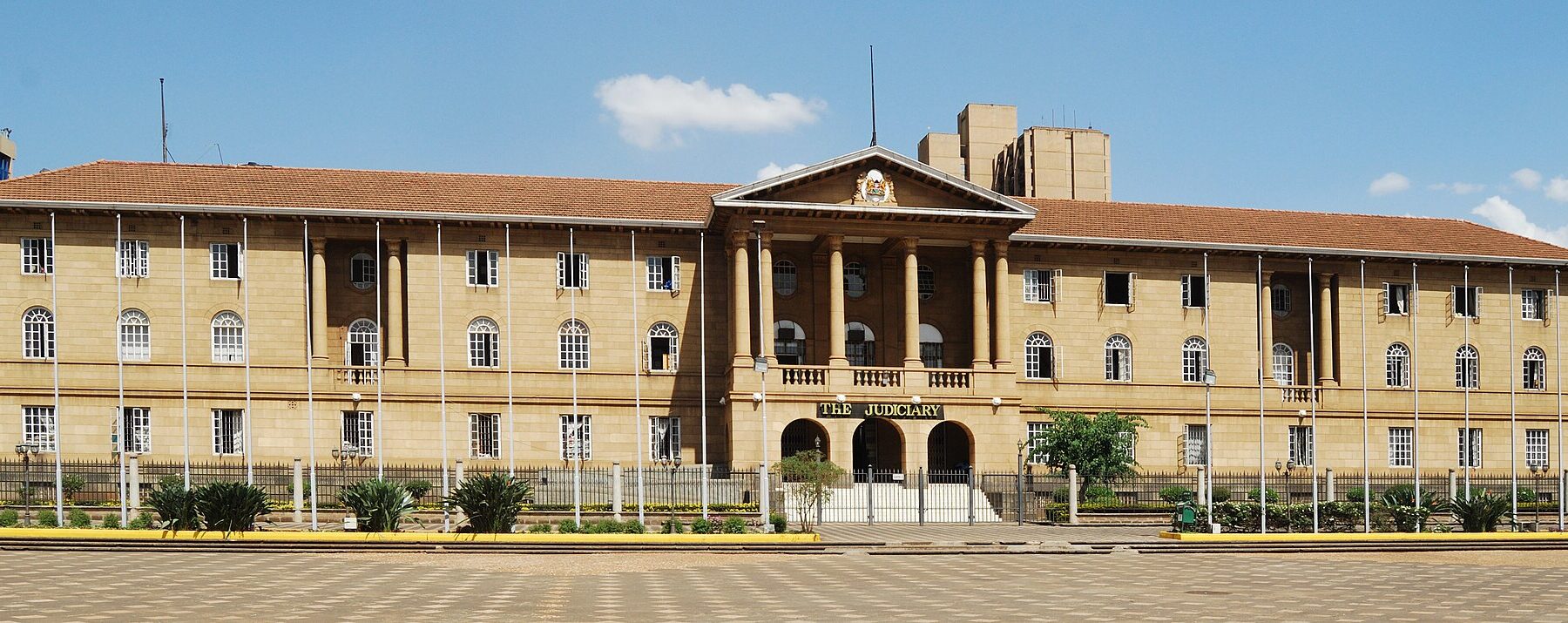 The colonnaded sandstone Judiciary building in Nairobi with a red-tiled roof, flagpoles and a paved forecourt under a blue sky