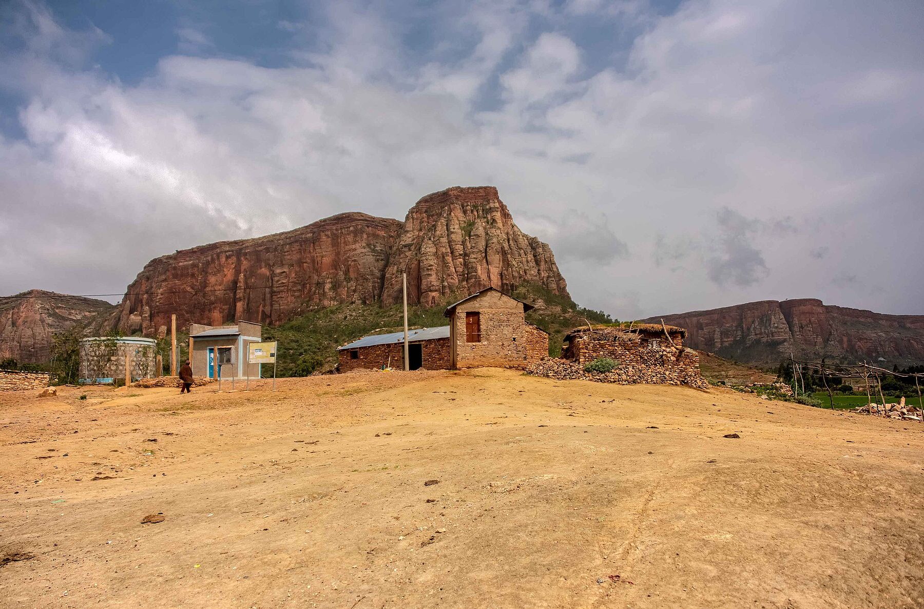 Red sandstone cliffs rise above a small village of stone buildings in rural Tigray under a cloudy sky