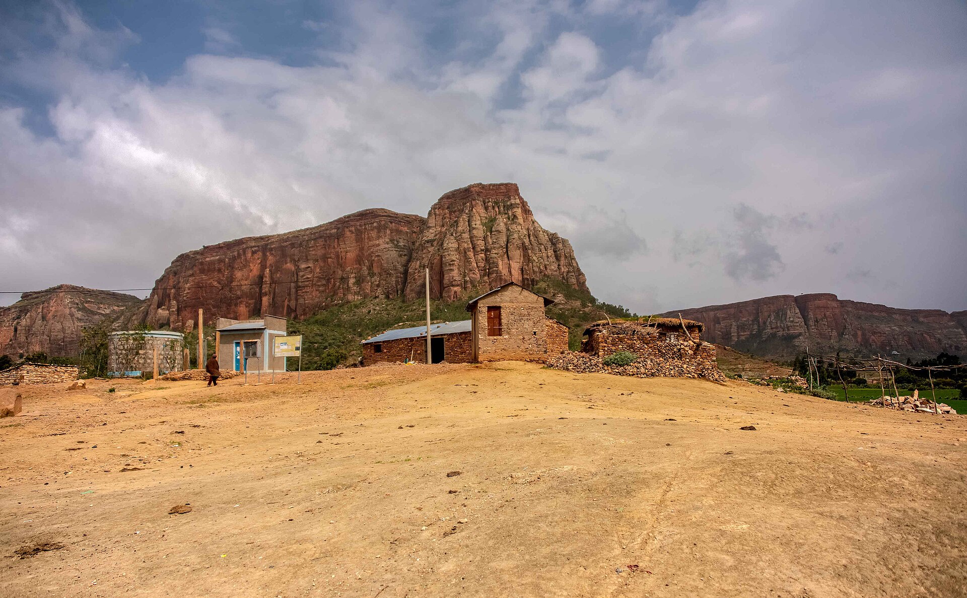 Red sandstone cliffs rise above a small village of stone buildings in rural Tigray under a cloudy sky