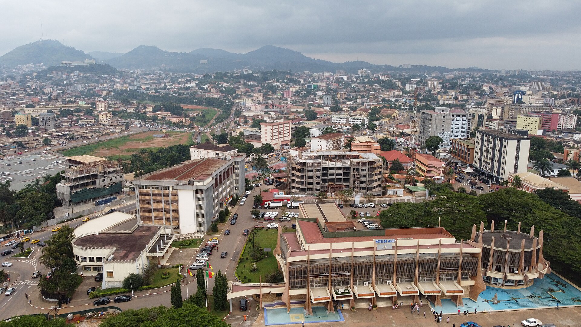 Aerial view of central Yaoundé with government and commercial buildings, tree-lined streets and green hills under an overcast sky
