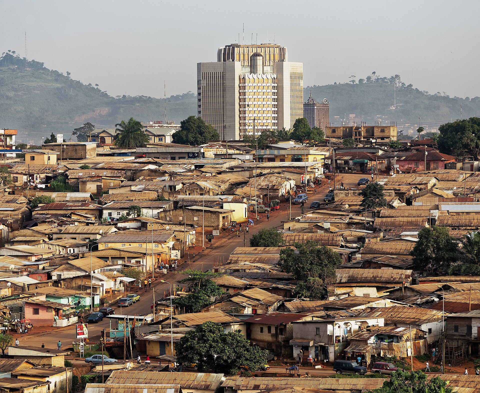 The tall headquarters tower of the Bank of Central African States rising above rows of low rusty-roofed houses in Yaoundé, with green hills behind