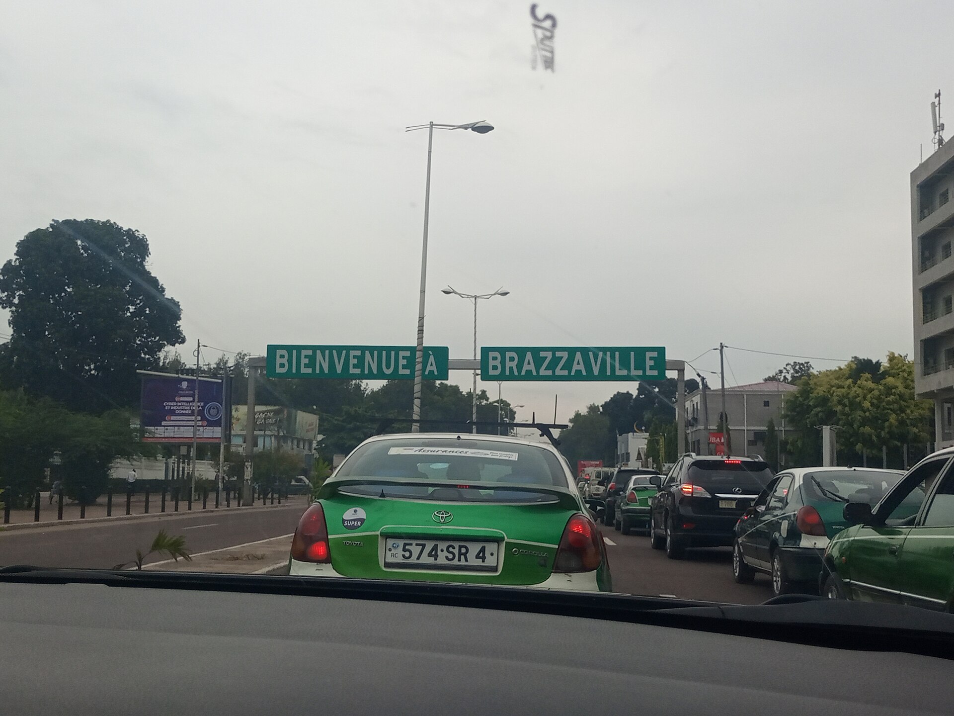 Cars in traffic on a Brazzaville street under a green road sign reading Bienvenue a Brazzaville, seen through a windscreen