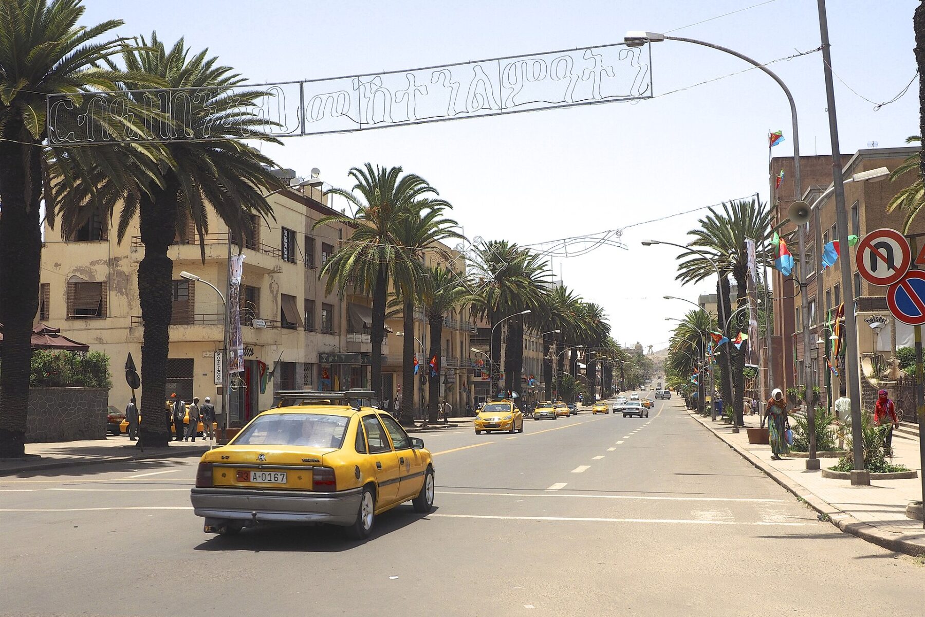 A wide street in Asmara lined with palm trees and low buildings, with a yellow taxi in the foreground