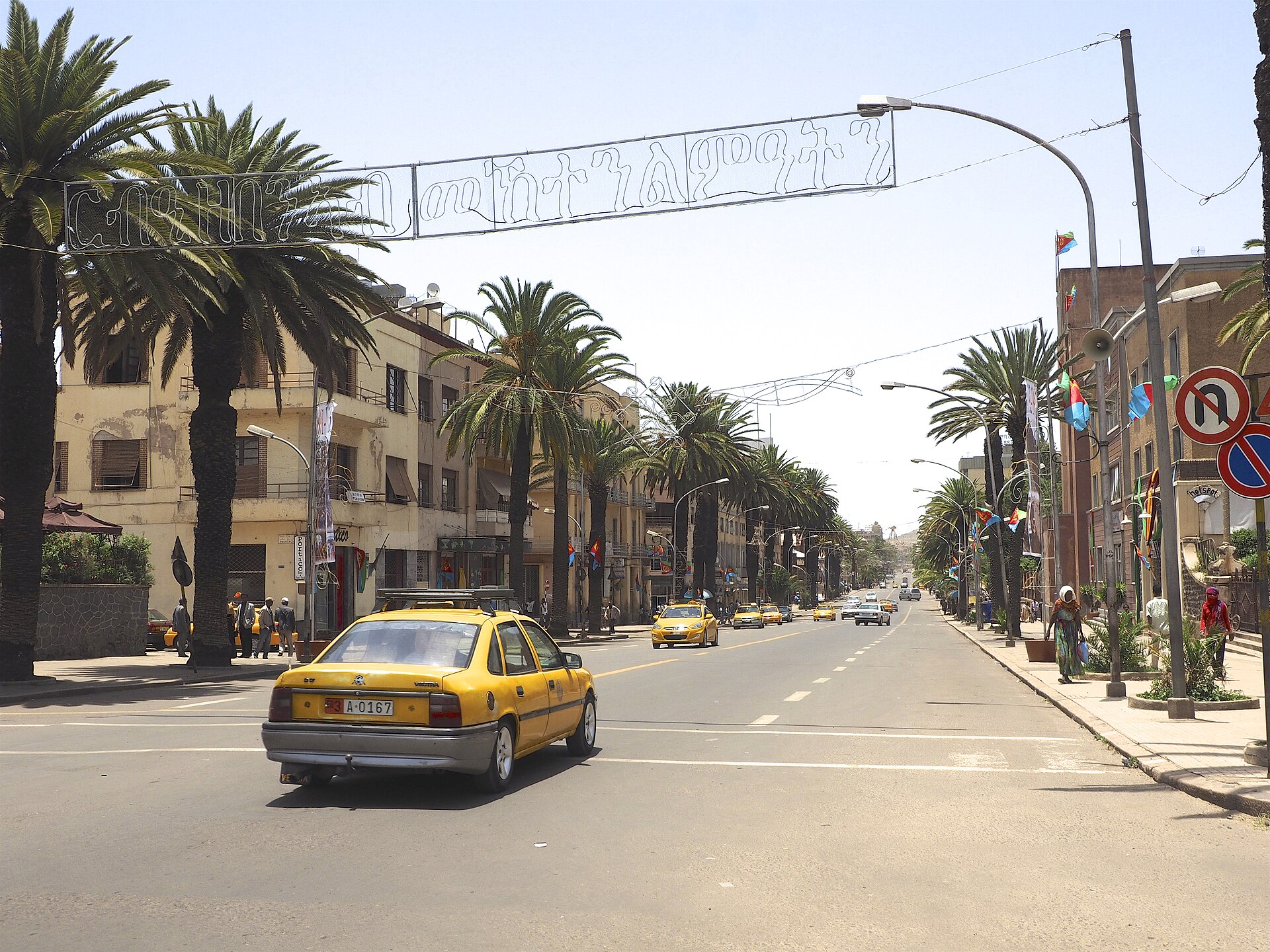 A wide street in Asmara lined with palm trees and low buildings, with a yellow taxi in the foreground