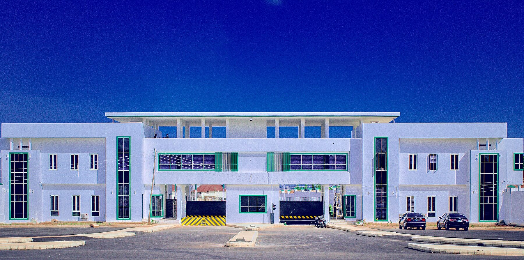 The white two-storey main entrance building of the Gombe State Government House with green window frames under a deep blue sky, two cars parked in front