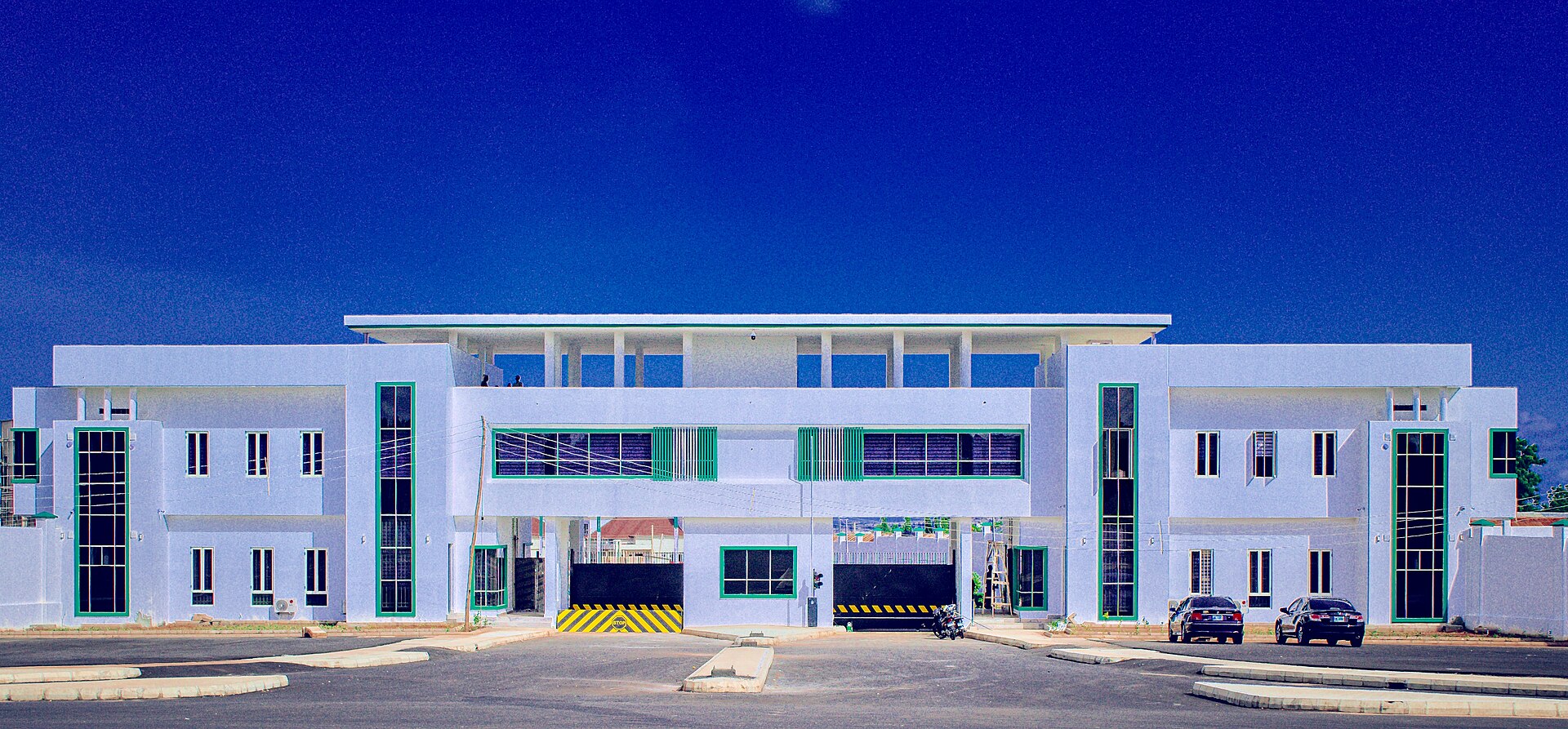 The white two-storey main entrance building of the Gombe State Government House with green window frames under a deep blue sky, two cars parked in front