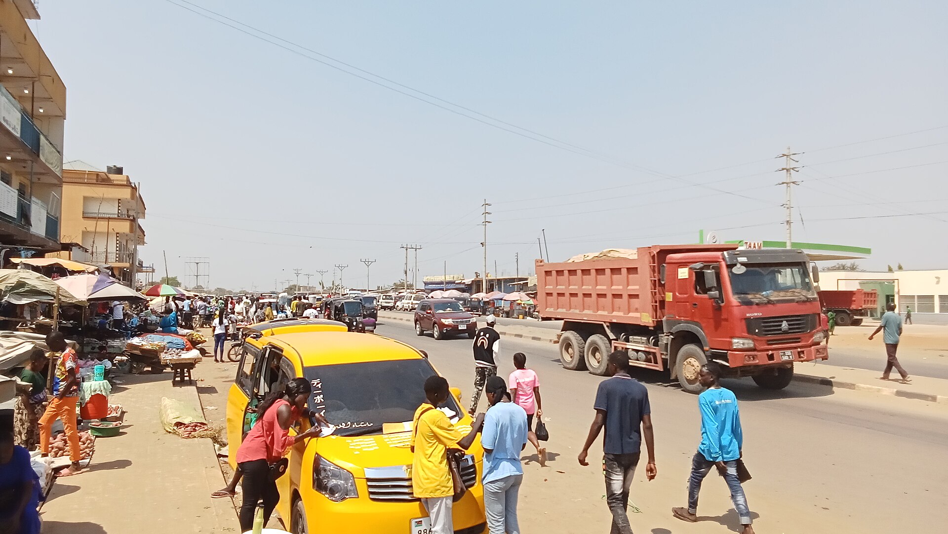 Pedestrians, a yellow taxi and a red tipper truck on a busy street beside market stalls in Gudele, Juba.