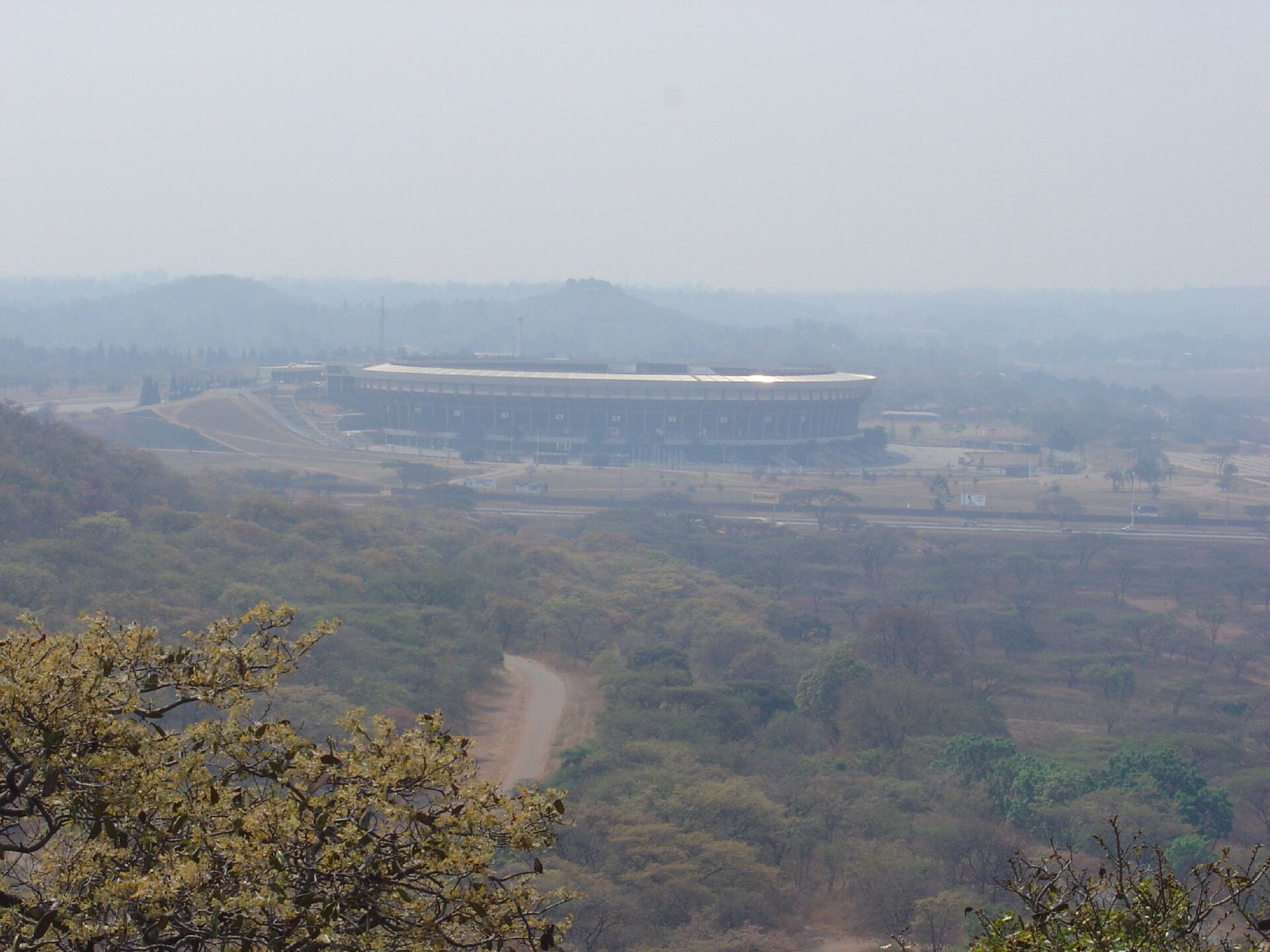 A large oval stadium on open ground below wooded hills, seen from a hillside in hazy light