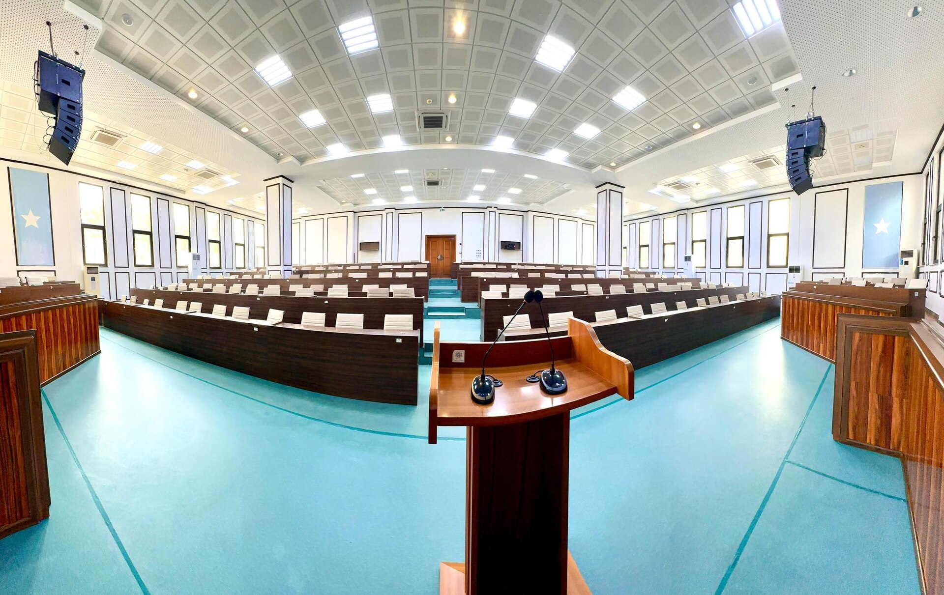 Empty parliamentary chamber with curved wooden benches, a lectern with microphones and blue banners with a white star
