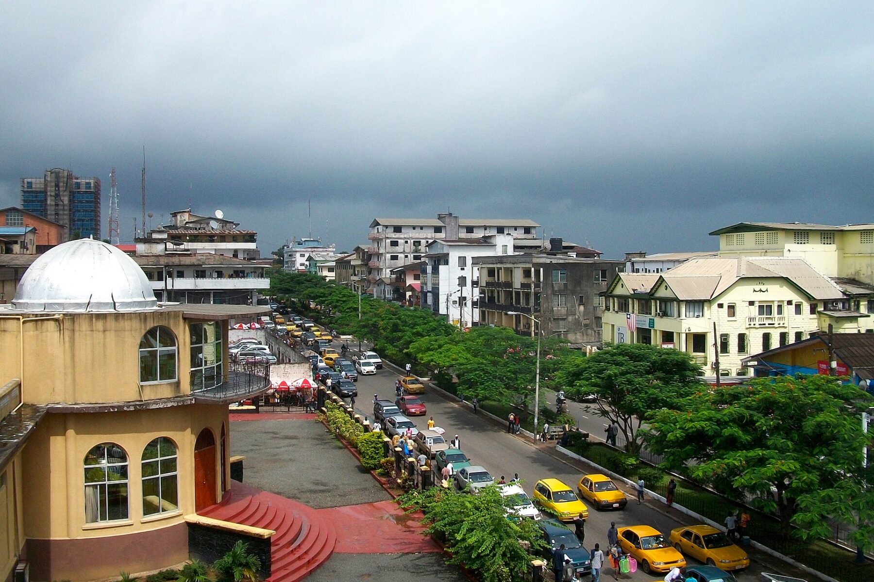 A tree-lined street in central Monrovia with yellow taxis, traffic and low buildings under a dark rain sky