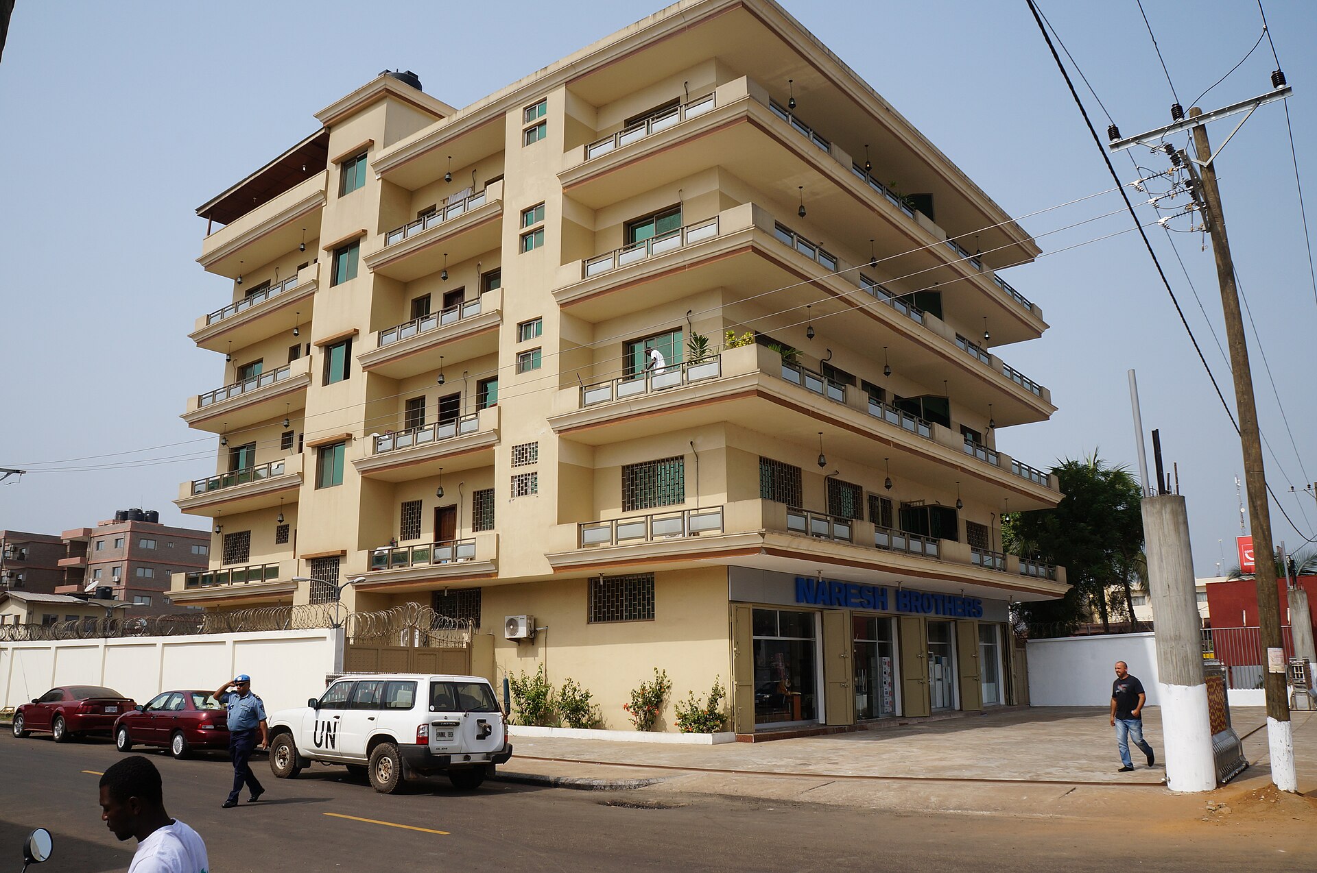A cream-coloured five-storey apartment building on a street corner in Monrovia, with a white UN vehicle and pedestrians