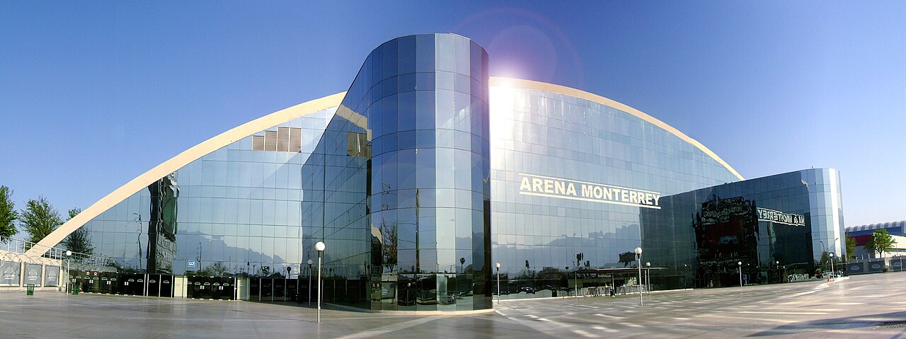 The curved glass front of Arena Monterrey under a clear blue sky, with the venue name across the facade.