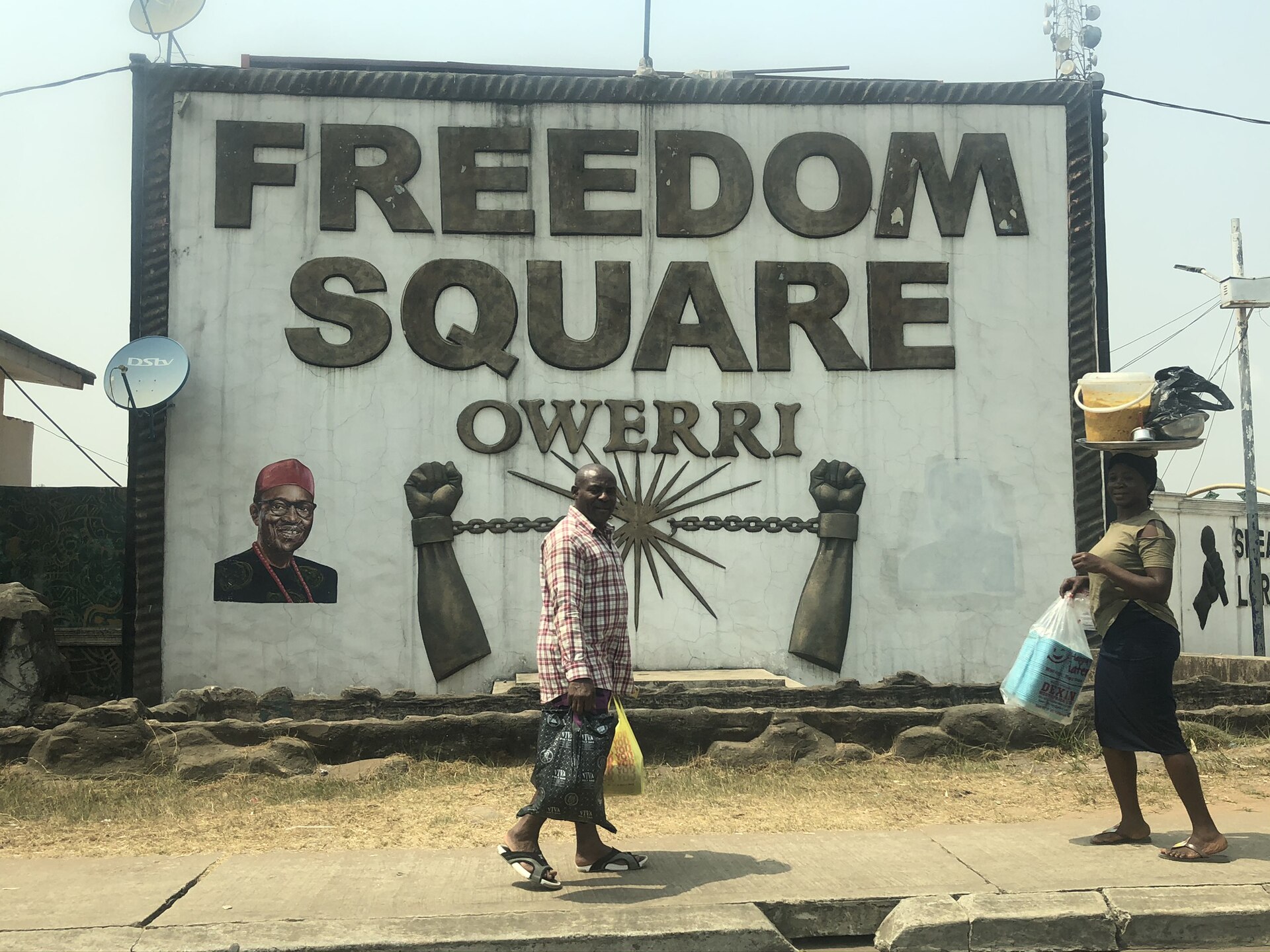 Freedom Square sign in Owerri with raised fists and a broken chain, as two pedestrians walk past