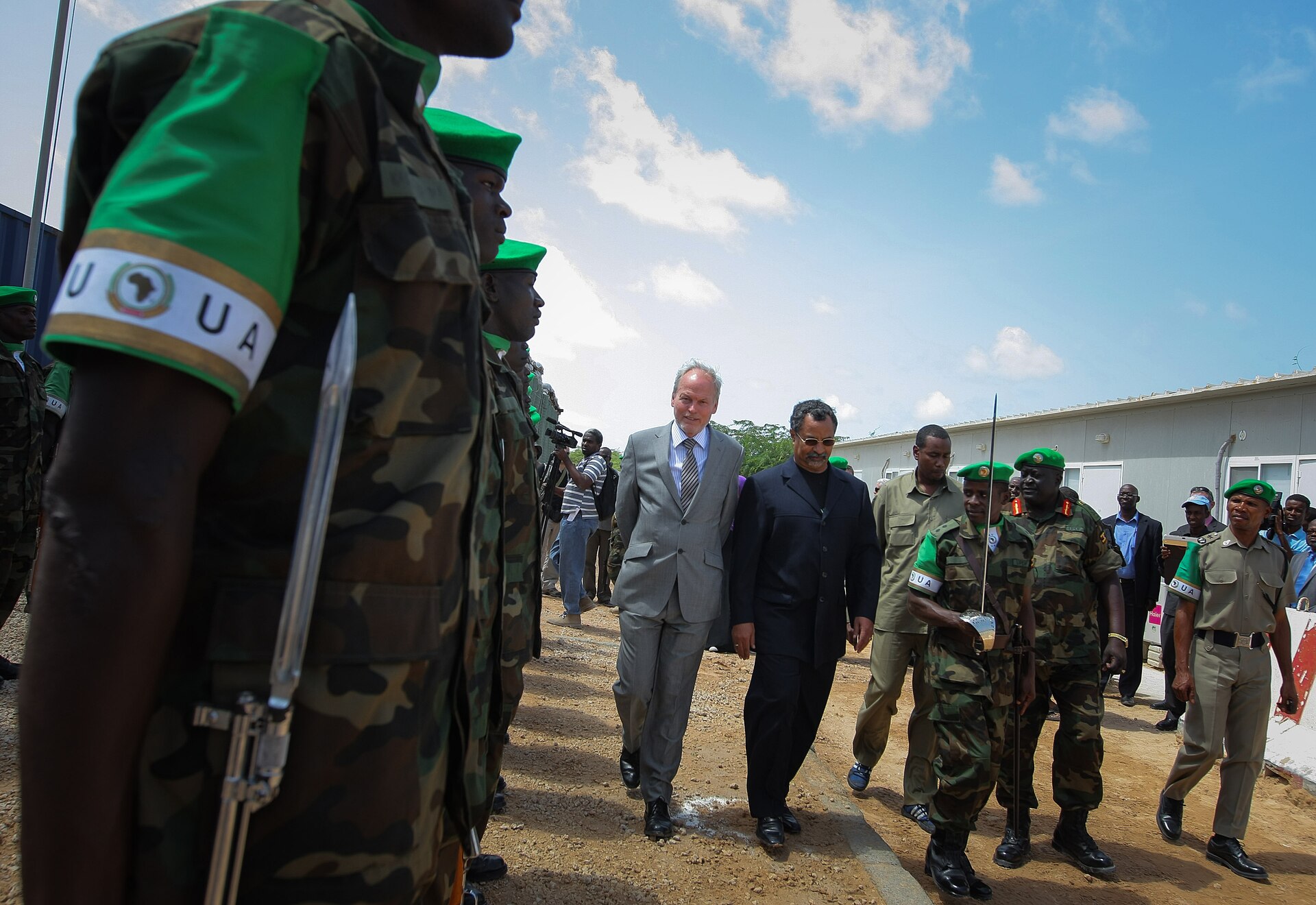 A man in a grey suit walks beside another man in dark clothes past a line of African Union soldiers in Mogadishu