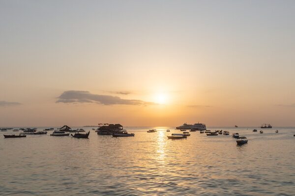 Small boats and a ferry on calm water in the harbour of Stone Town, Zanzibar, at sunset, with a quay and low buildings on the left