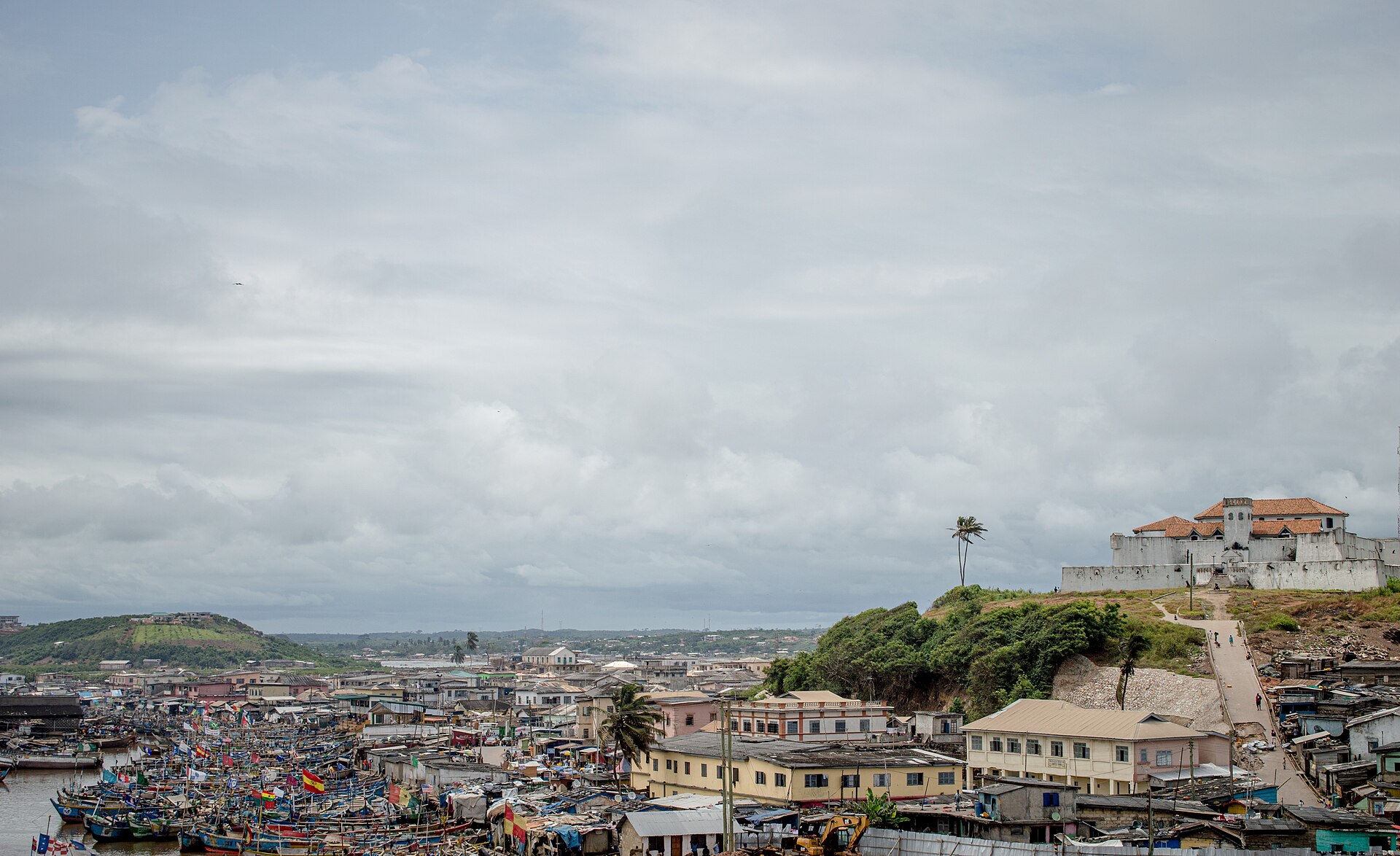A crowded fishing harbour with colourful boats below a white fort on a hill under a cloudy sky
