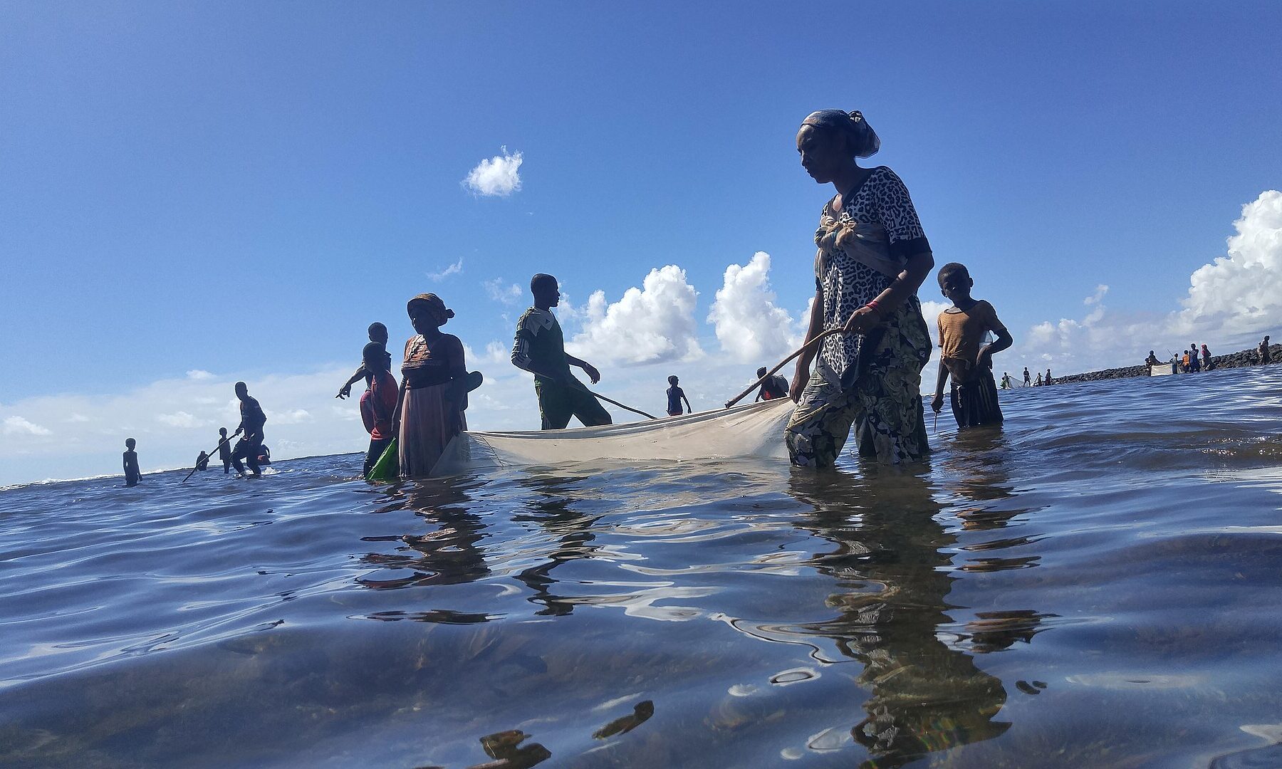 Women and children wade in shallow sea water pulling a fishing net off the coast of a Comoros island.