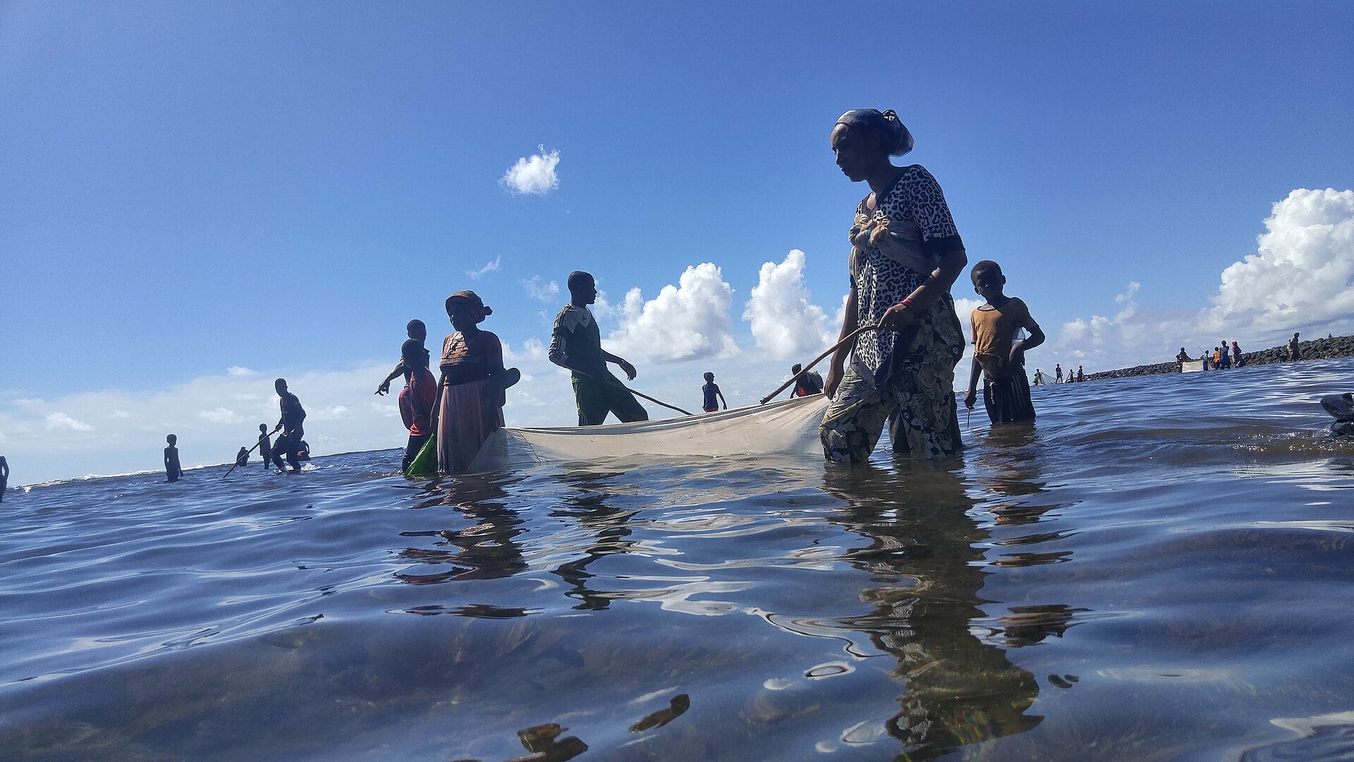 Women and children wade in shallow sea water pulling a fishing net off the coast of a Comoros island.