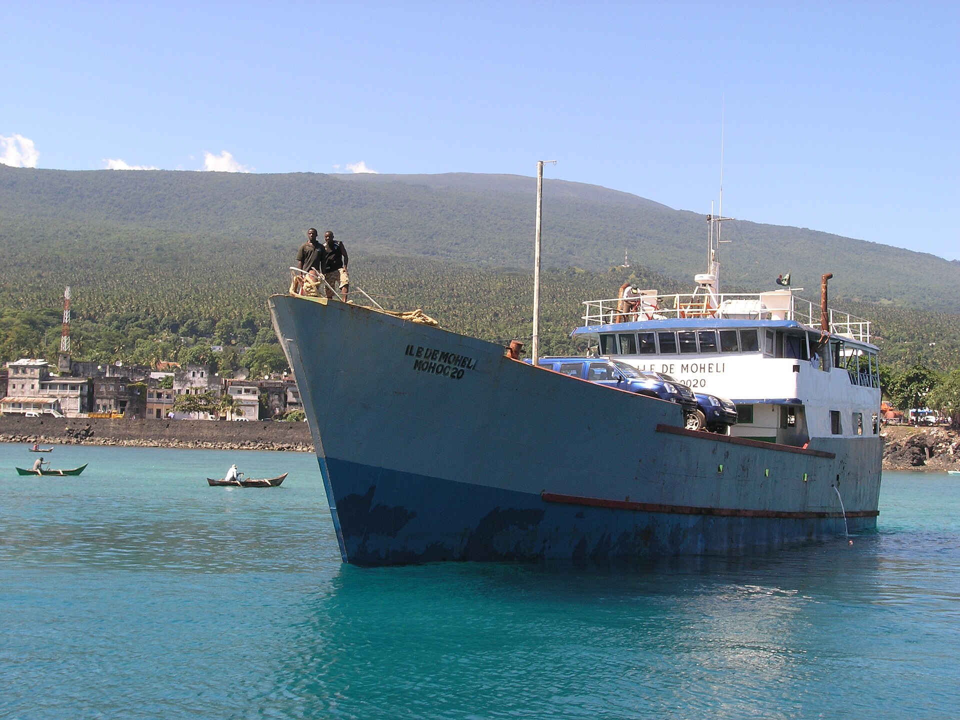 A blue cargo and passenger ship carrying cars on deck sits in turquoise water off Fomboni, Moheli, with small canoes, harbour buildings and green hills behind.
