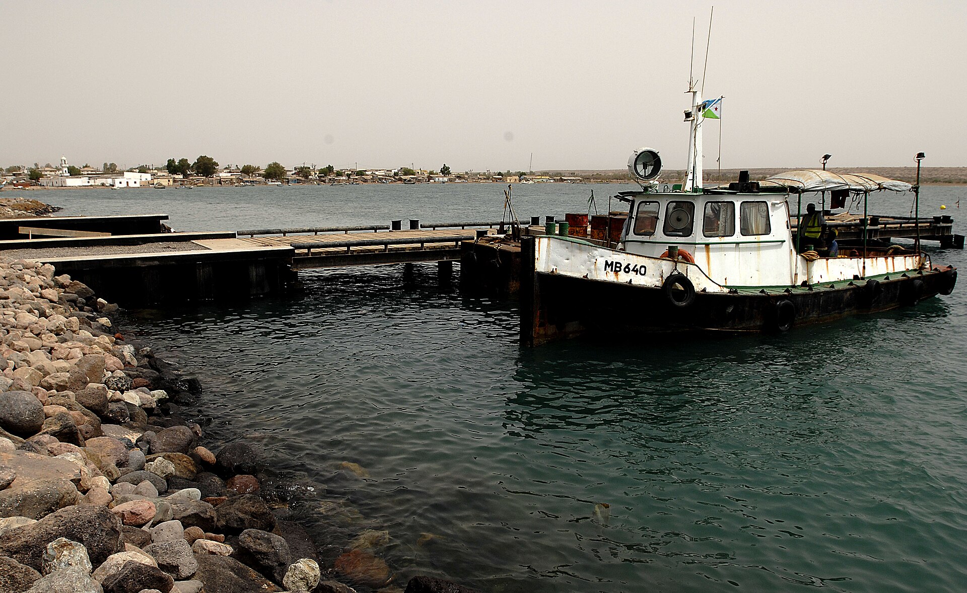 A white and black tugboat flying the Djibouti flag moored at a pier in Obock harbour, with rocks in the foreground and the low town on the far shore
