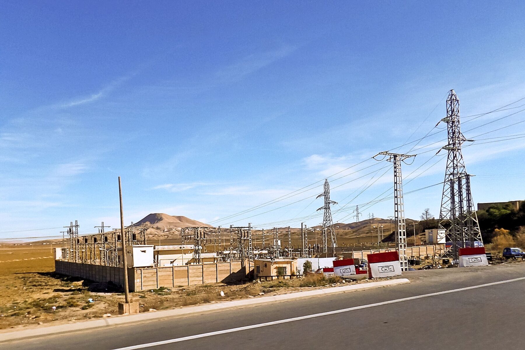 A roadside electrical substation with pylons and transmission lines on dry plains in Algeria, with red and white GRTE blocks by the road