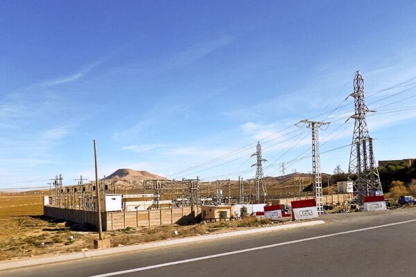 A roadside electrical substation with pylons and transmission lines on dry plains in Algeria, with red and white GRTE blocks by the road
