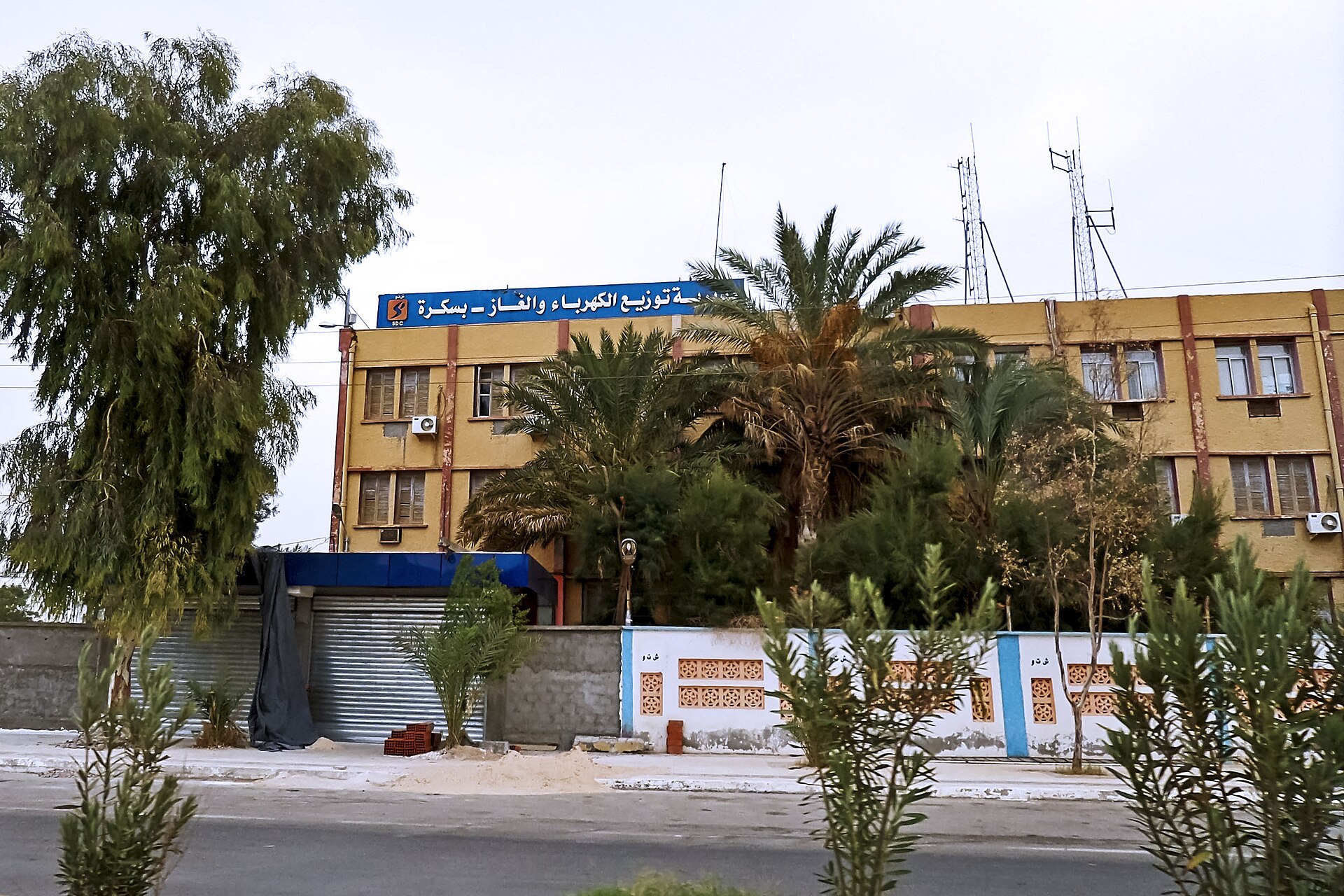 A yellow three-storey Sonelgaz electricity and gas distribution office in Biskra behind palm trees, with an Arabic sign on the roof