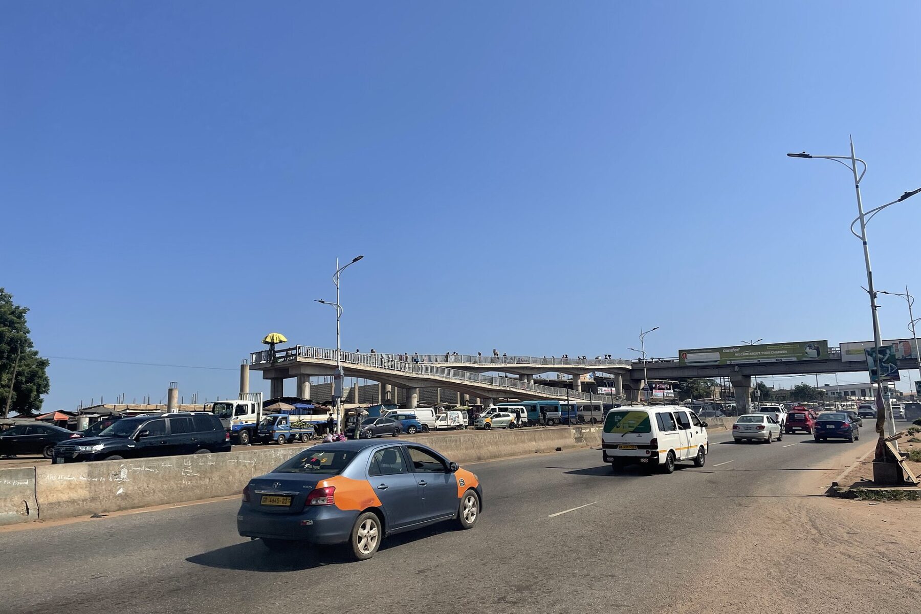 Cars and a minibus on a wide road beneath the Madina footbridge in Accra under a clear blue sky
