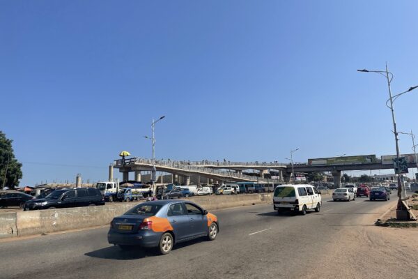 Cars and a minibus on a wide road beneath the Madina footbridge in Accra under a clear blue sky