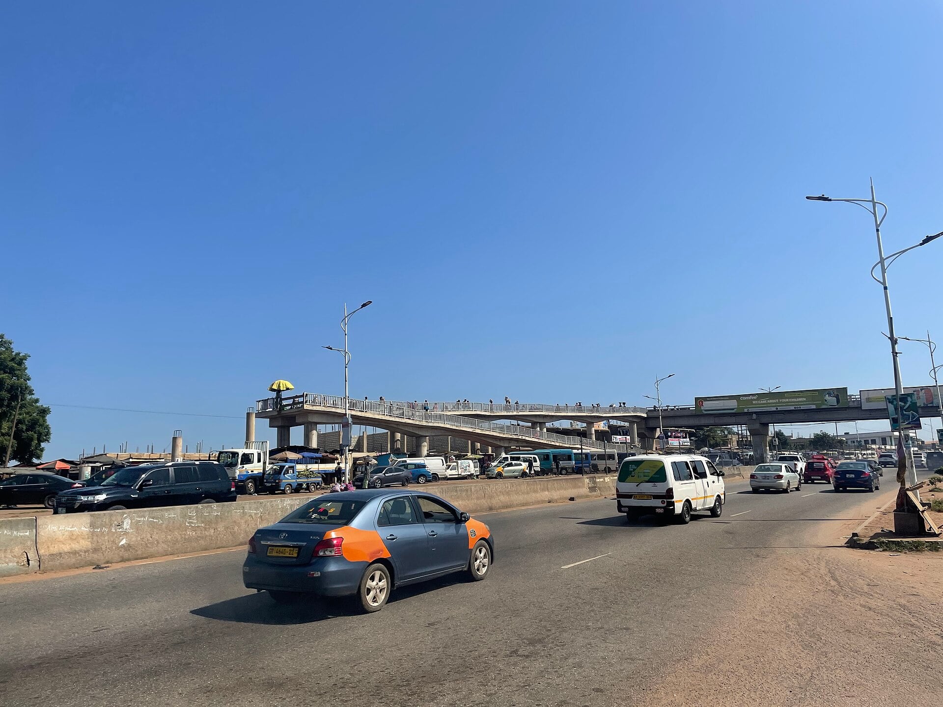 Cars and a minibus on a wide road beneath the Madina footbridge in Accra under a clear blue sky