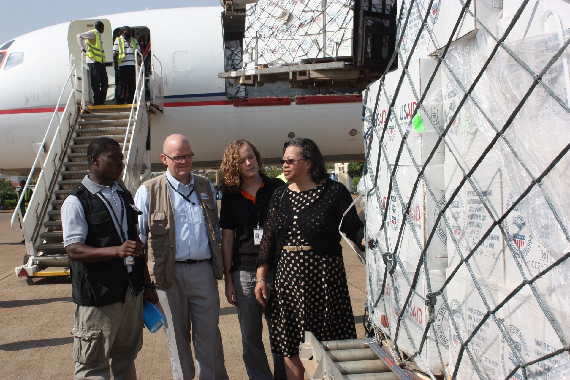 Aid officials stand beside pallets of boxed relief supplies marked USAID next to a cargo plane on an airport apron in South Sudan.