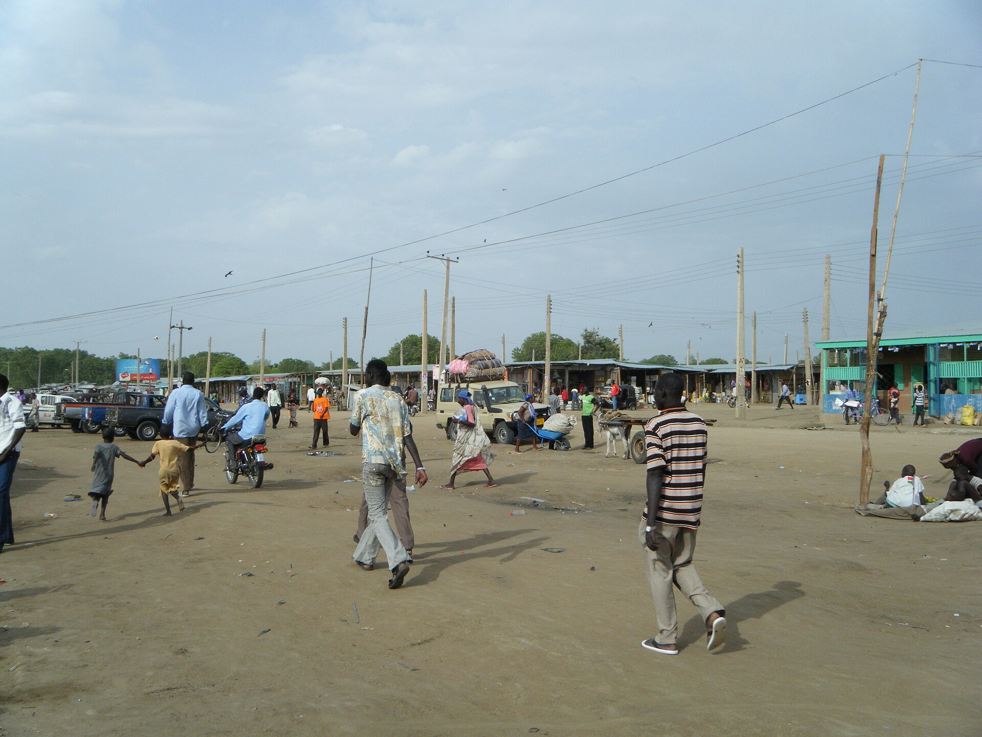 People walk across a wide dusty square in front of a row of market stalls under a cloudy sky in Bor, South Sudan.