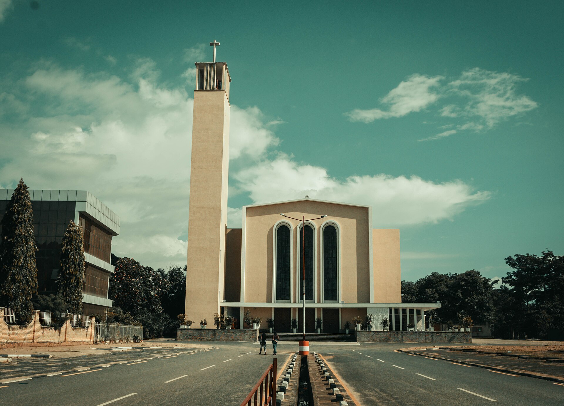 A cream-coloured cathedral with a tall square bell tower topped by a cross stands at the end of a wide divided road, with two people walking and trees on either side.